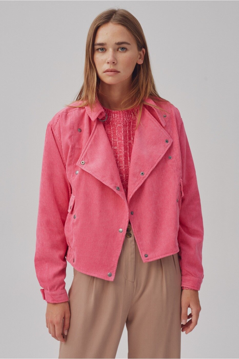 A woman poses in a pink corduroy jacket and tan pants against a neutral backdrop, showcasing a stylish and modern fashion look.