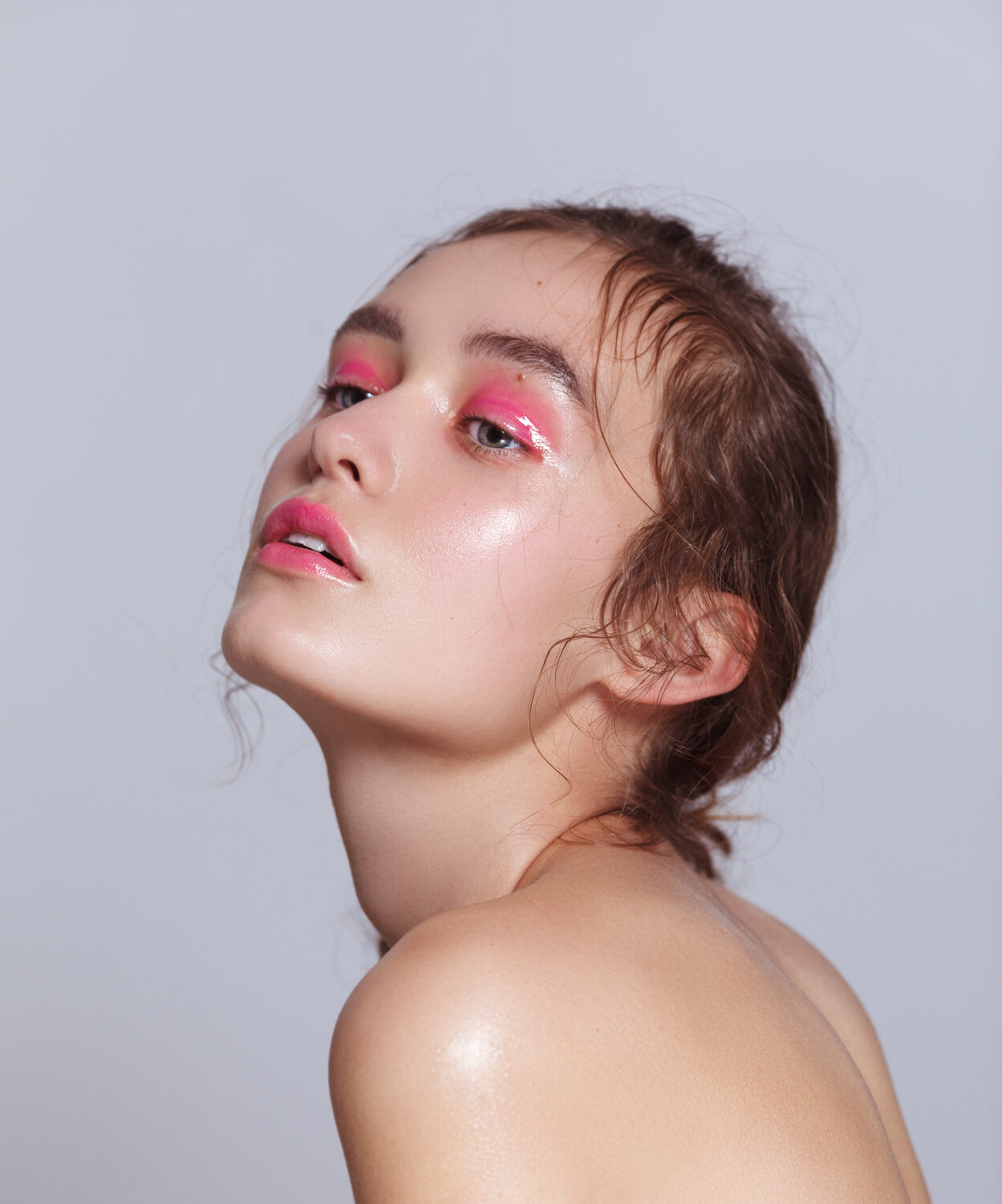 Close-up beauty shot of a young fair-skinned woman with pink glossy eyeshadow and lips, wet hair, and bare shoulders against a grey background.