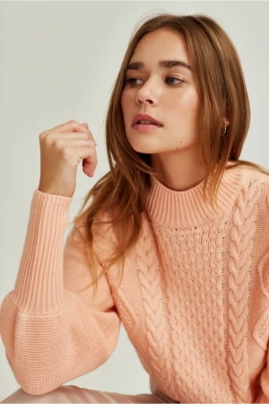 Close-up portrait of a woman in her 20s with long brown hair wearing a pink cable-knit sweater against a plain light background