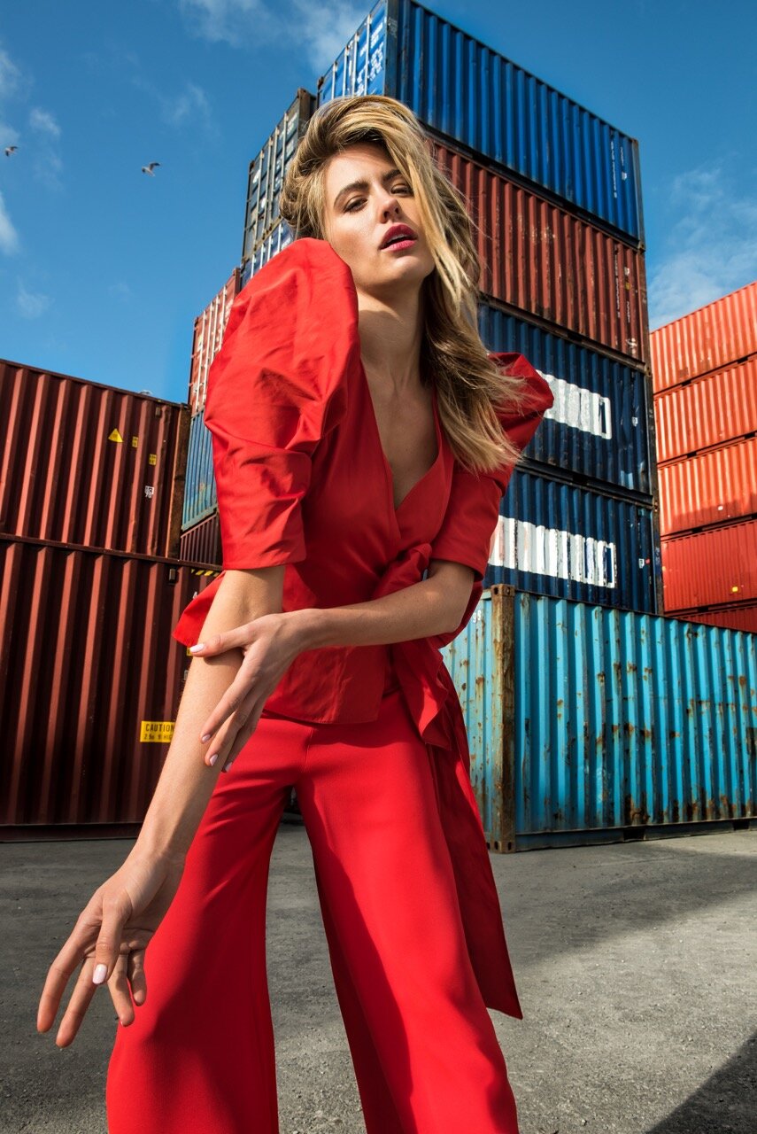 A stylish woman in a red outfit poses in front of stacked shipping containers under a bright blue sky, showcasing a fashion editorial.