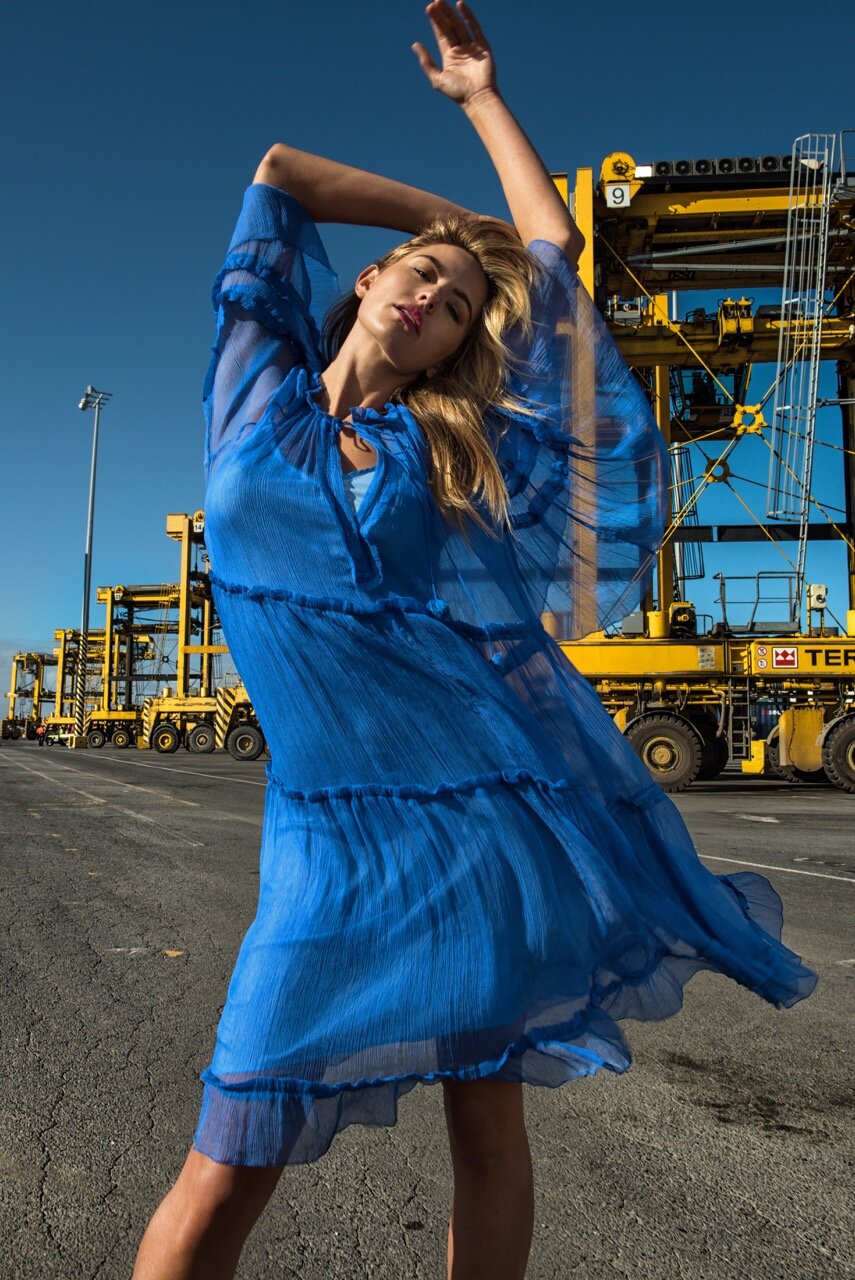 A blonde woman in a flowing blue dress poses with arms raised at an industrial port with shipping cranes under a clear blue sky.