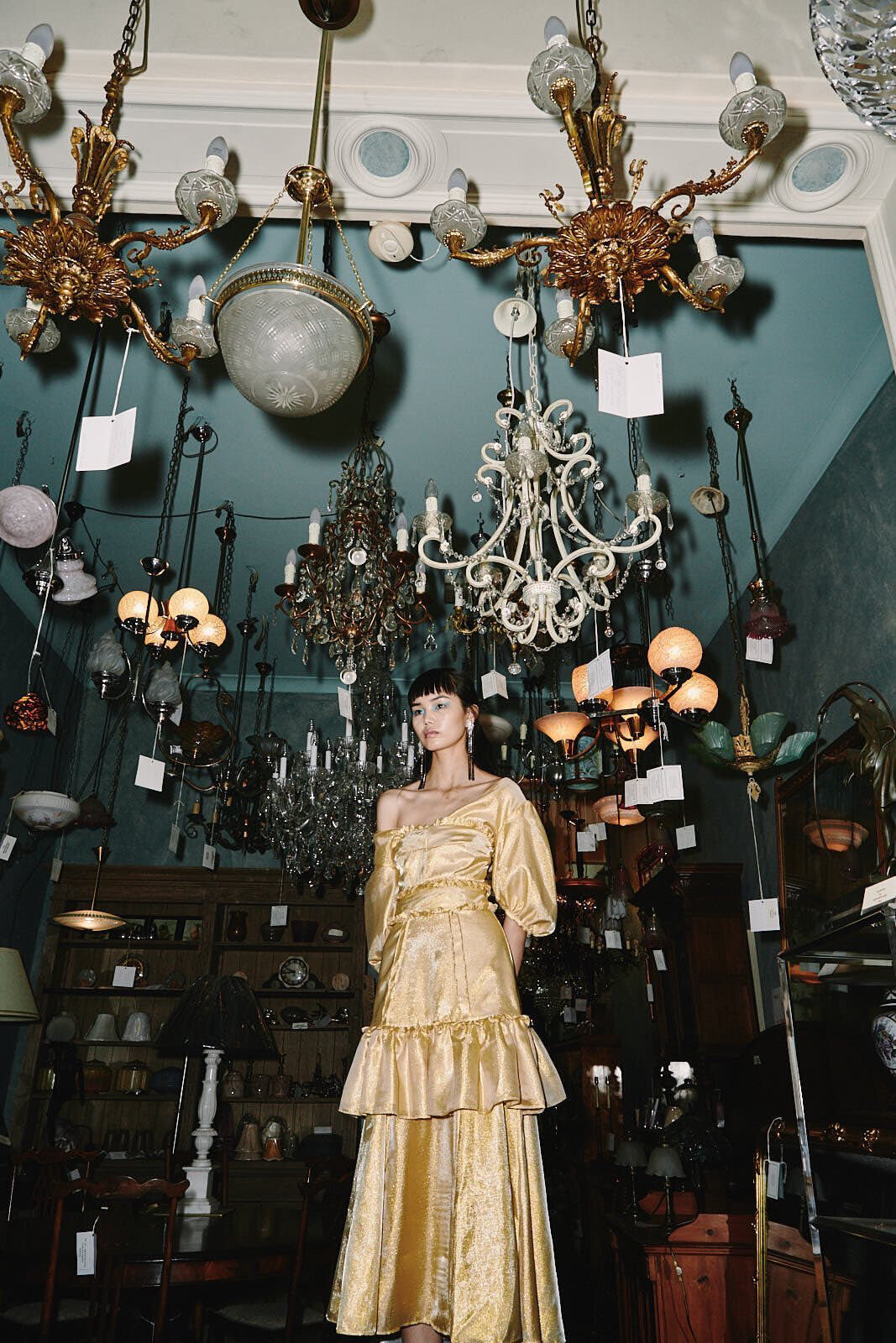 A woman poses in a golden dress in an antique store filled with chandeliers, showcasing vintage fashion and a nostalgic atmosphere.