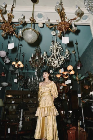 A woman poses in a golden dress in an antique store filled with chandeliers, showcasing vintage fashion and a nostalgic atmosphere.