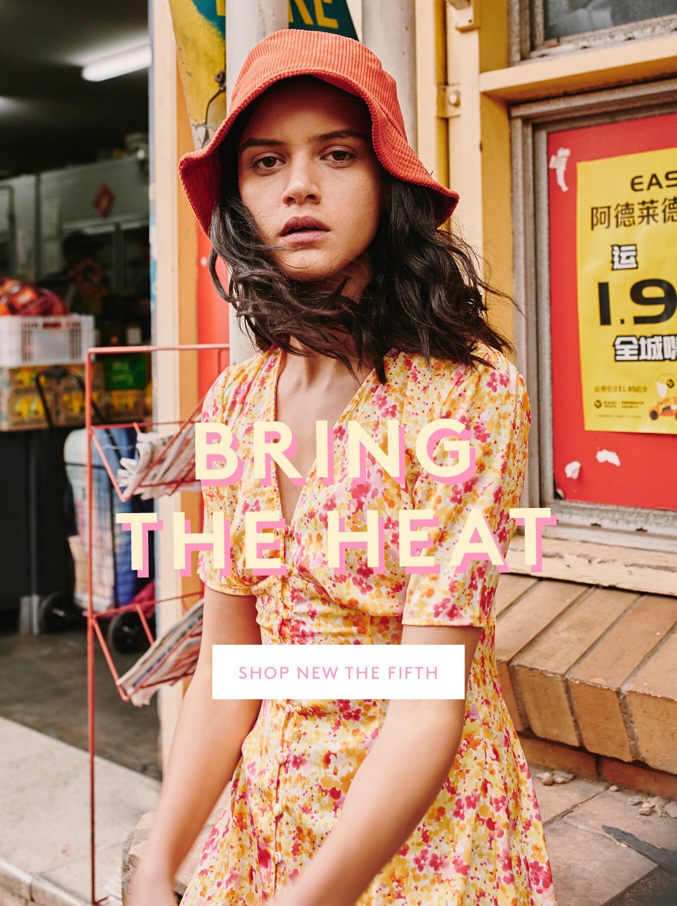 A fashionable young adult female in a floral dress and orange hat poses outdoors near a colorful, slightly weathered building.