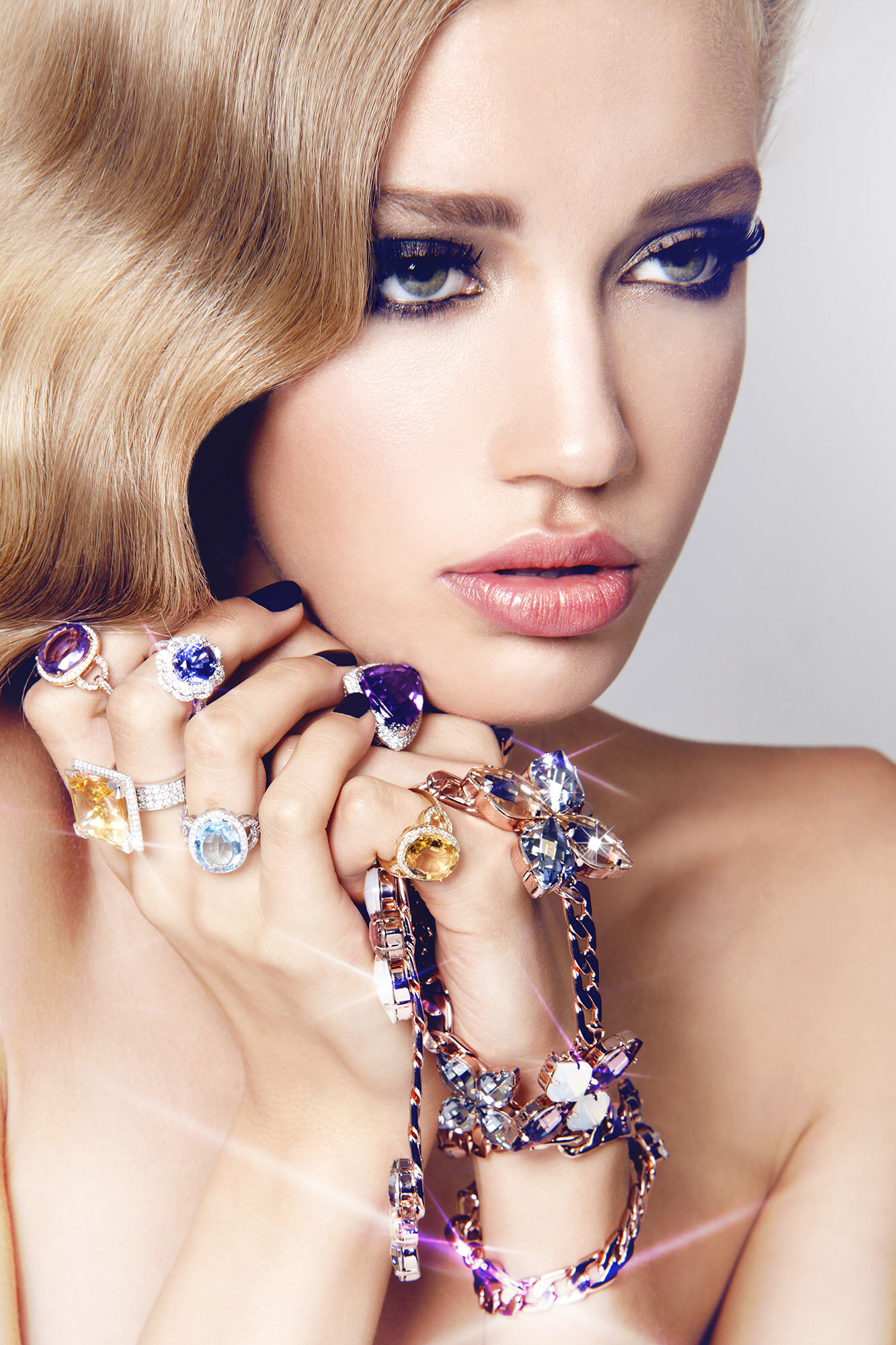 A blonde woman with makeup is posing and holding colorful rings and a jeweled bracelet in a close-up studio fashion shot.