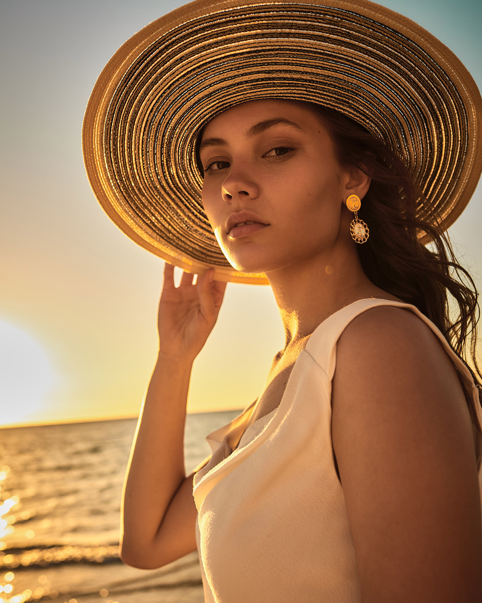 Woman in sun hat and cream dress poses on beach during sunset. Warm golden tones, ocean backdrop. Fashion and lifestyle concept.