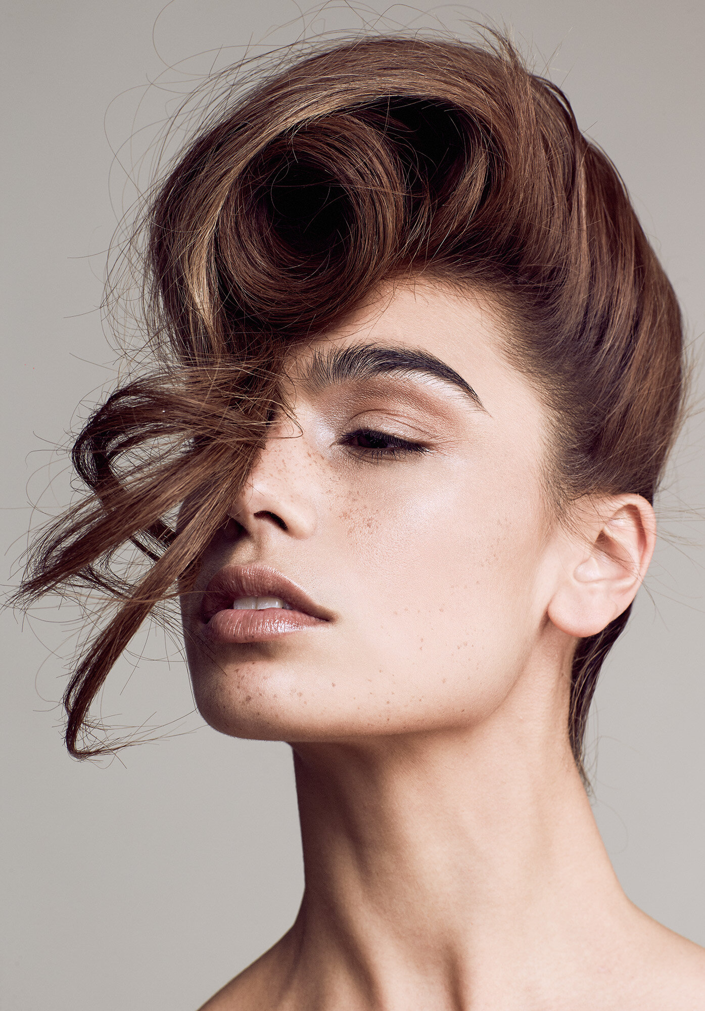 Close-up of a brunette adult female with freckles, an elaborate curled hairstyle, glossy lips, and fair skin, captured in a studio beauty shot.
