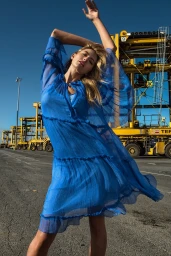 Model wearing a sheer blue dress posing with arms raised against an industrial dock background with large yellow cranes