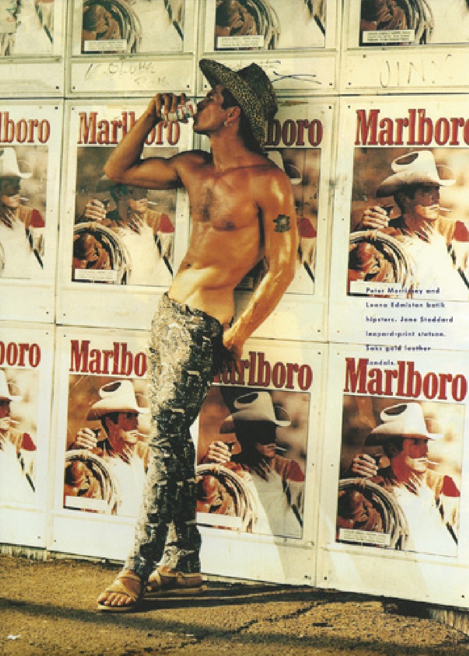 Shirtless man in a cowboy hat drinks from a can, leaning against a wall of Marlboro ads in an outdoor setting, tan sandals.