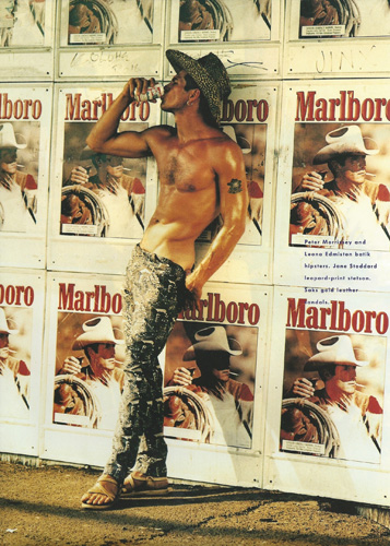 Shirtless man wearing a cowboy hat and patterned trousers leaning against a wall covered in Marlboro posters