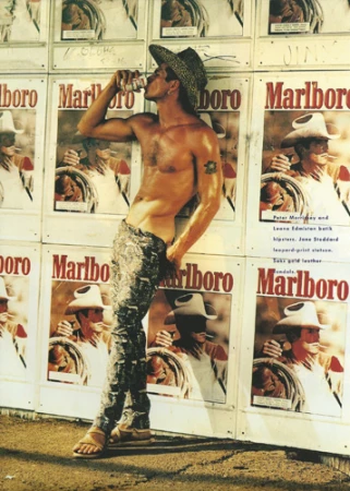 Shirtless man wearing a cowboy hat and patterned trousers leaning against a wall covered in Marlboro posters