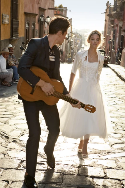 A man in a suit plays guitar for a woman in a wedding dress on a sunny cobblestone street, with blurred figures in the background.