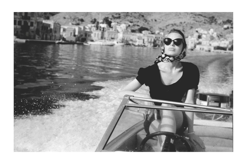 Black-and-white portrait of a woman in sunglasses and headscarf at the helm of a motorboat on the water near a hillside town.