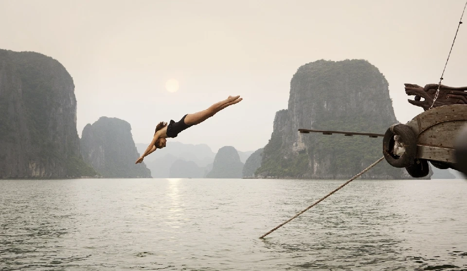 Woman in black swimsuit dives into ocean off boat; limestone karsts and misty sky in background; Travel photography in Ha Long Bay, Vietnam.