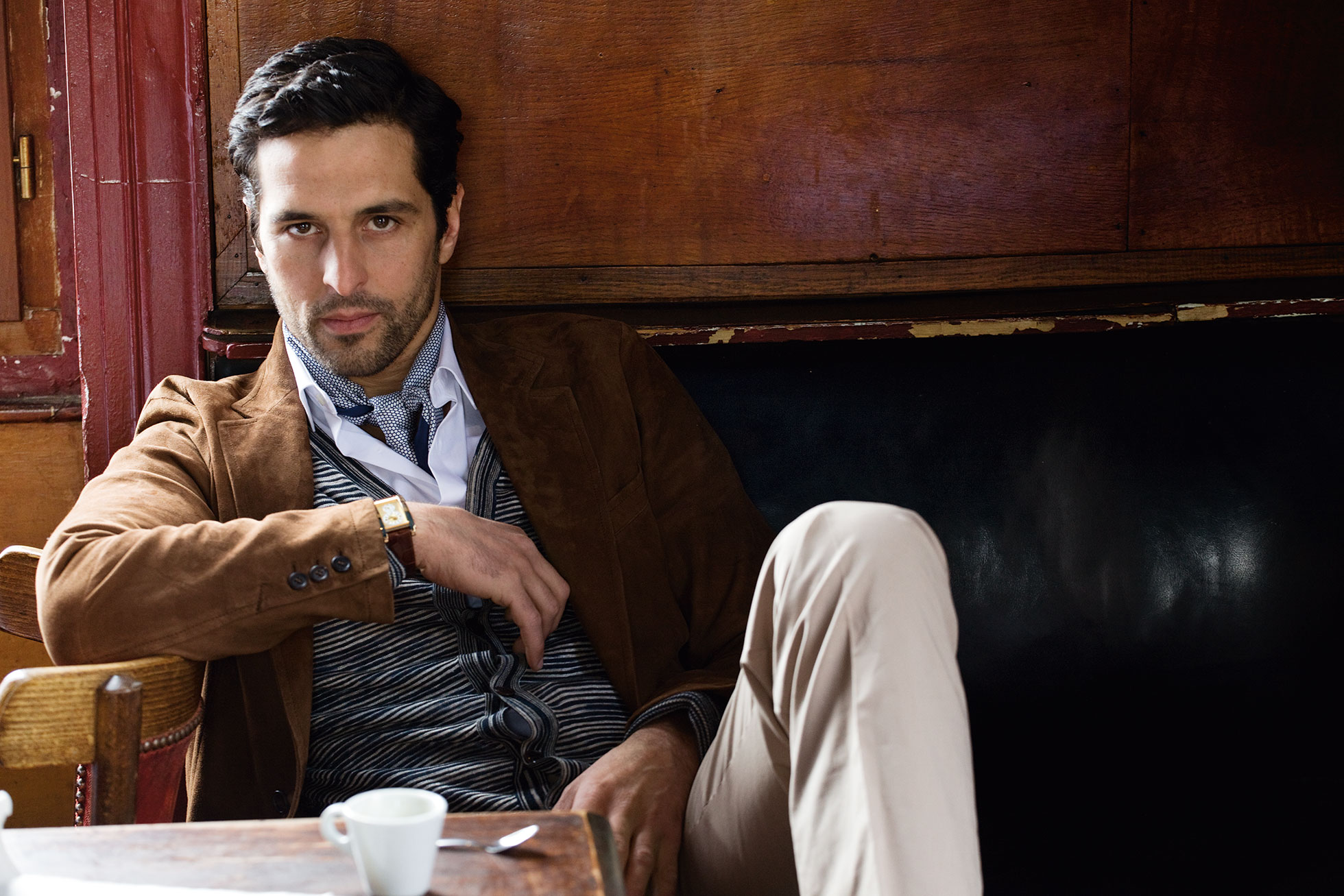 A man in a brown jacket sits at a cafe table with a cup of coffee, posing indoors in a classic and sophisticated fashion style.