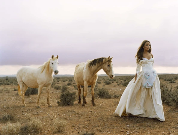An adult female model stands in a bridal gown in a dry field next to two horses beneath a cloudy sky at dusk in a fashion editorial.