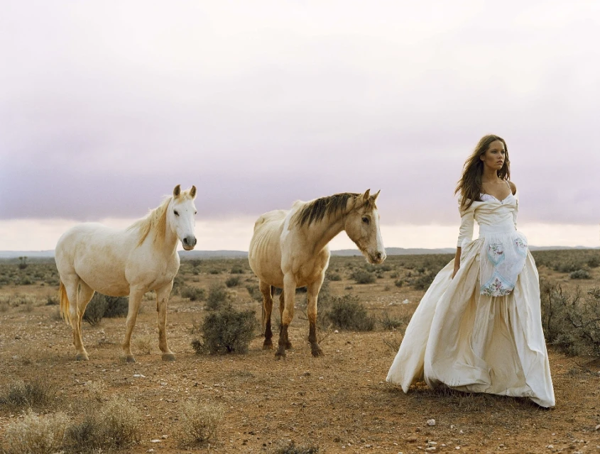 Woman in an elegant dress stands in a desert landscape alongside two horses under a pastel sky, fashion photography in an outdoor setting.