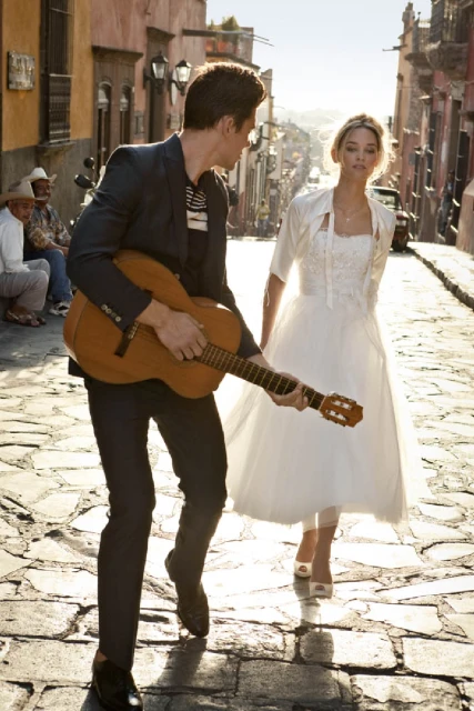 A man plays guitar while a woman in a white dress stands on a cobblestone street with old buildings, showcasing outdoor fashion.