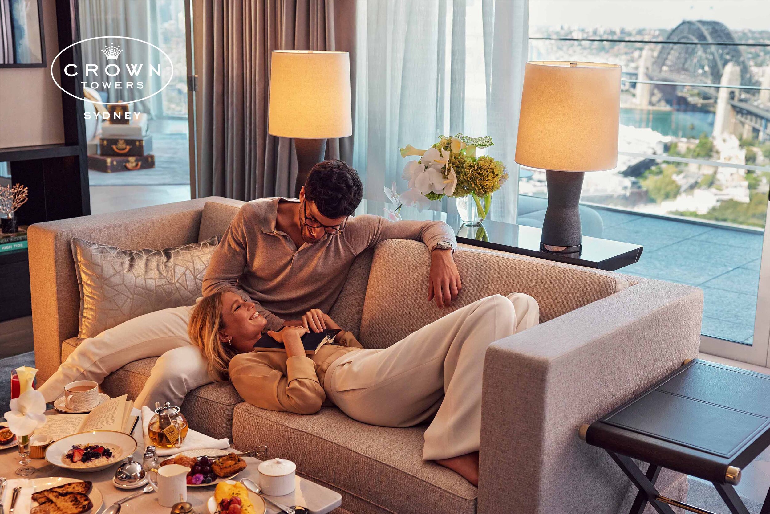 Couple relaxing on a neutral couch in a modern apartment setting with views of a cityscape and the Sydney Harbour Bridge.