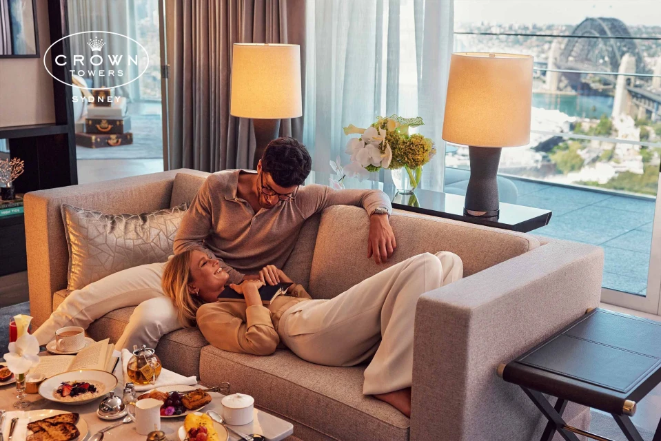 Couple relaxing on a neutral couch in a modern apartment setting with views of a cityscape and the Sydney Harbour Bridge.