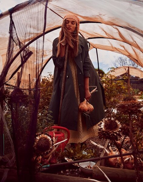 Woman in a coat and headscarf stands in a plant-filled greenhouse, holding produce, with natural light filtering through.