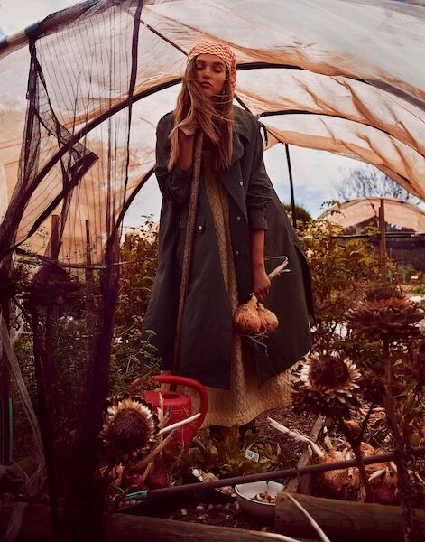 Woman in a coat and headscarf stands in a plant-filled greenhouse, holding produce, with natural light filtering through.