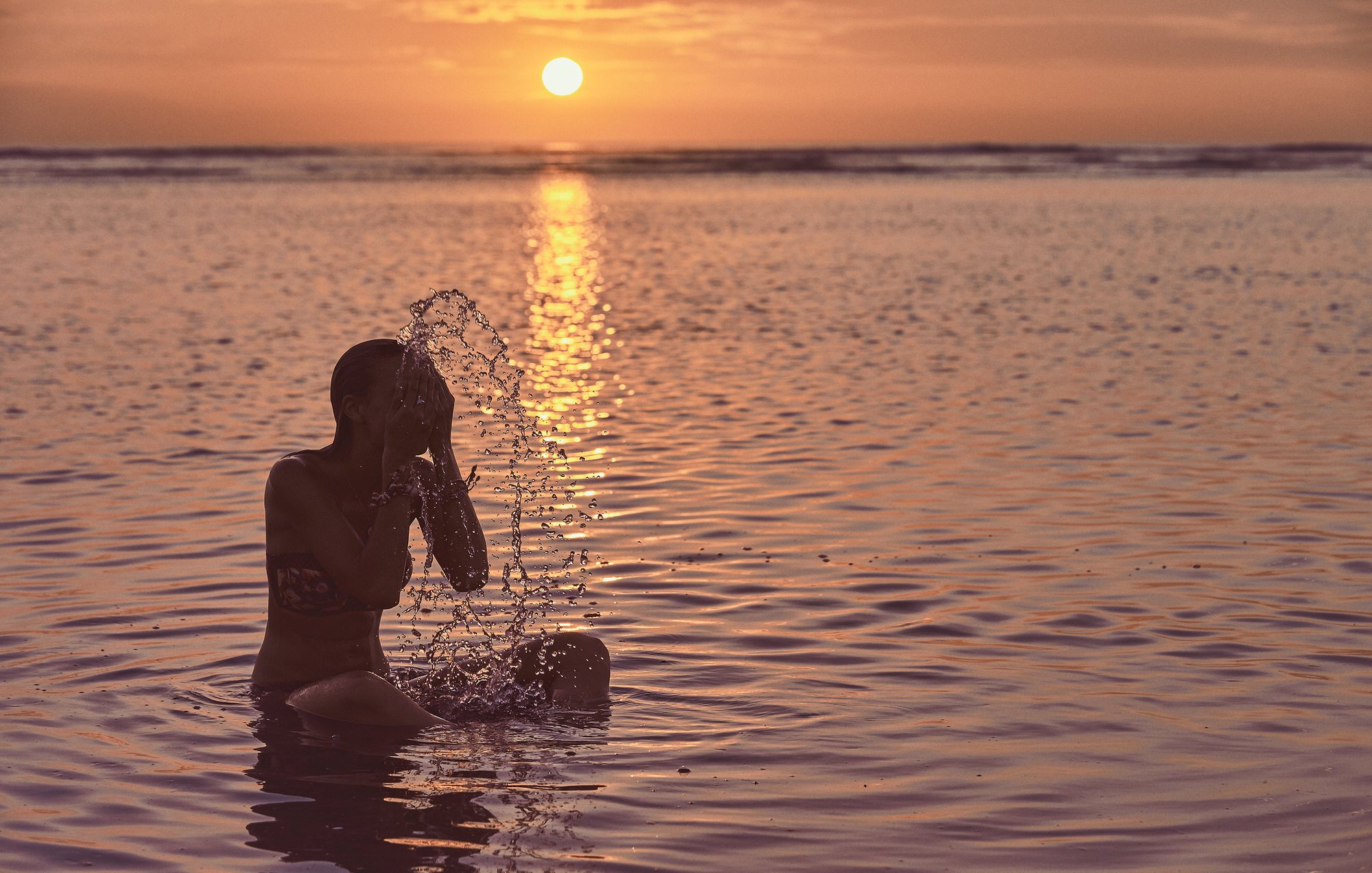Woman seated in shallow ocean water at sunset, splashing water over her face with the sun reflecting on the sea.