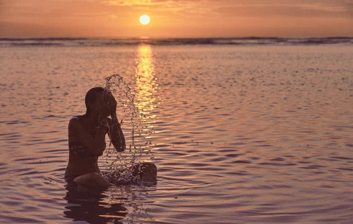 Woman seated in shallow ocean water at sunset, splashing water over her face with the sun reflecting on the sea.