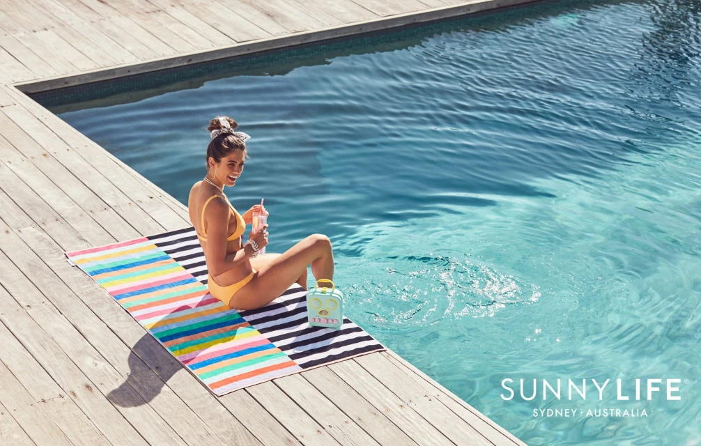 A woman in a yellow bikini sitting on a colorful striped towel next to a pool with a drink. Sunny day by the pool, summertime.