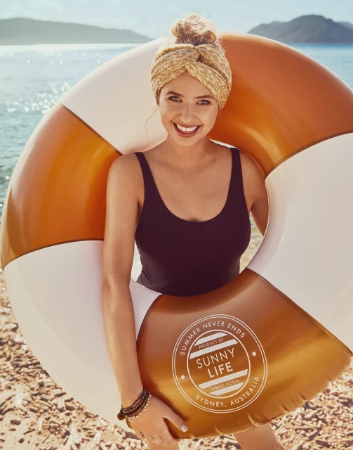 Woman in black swimsuit holds a gold and white inflatable ring, smiling on a sunny beach with ocean and headscarf.