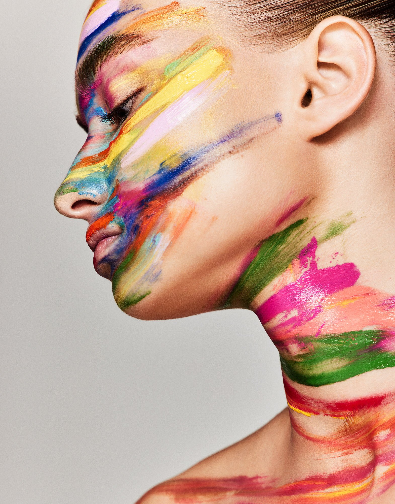 Close-up of a young woman's face and upper body covered in bright multi-colored paint in a beauty or fashion shot. Indoor studio.