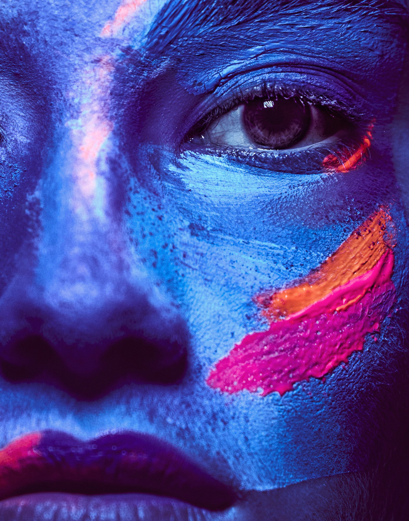 Close-up of a person's face covered in blue body paint, with pink and orange streaks across the cheek and brow, illuminated by vibrant lighting.