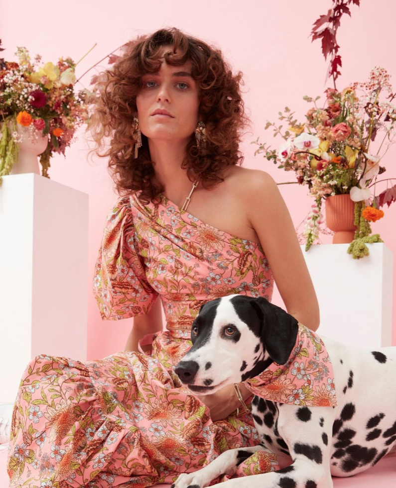 Woman in a floral one-shoulder dress poses with a Dalmatian dog in a matching floral bandana, surrounded by floral arrangements on pink.