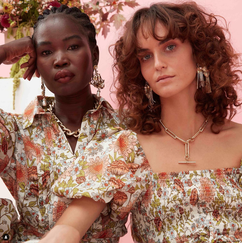 Two women pose in floral dresses with gold jewelry against a pink background, showcasing fashion and beauty.