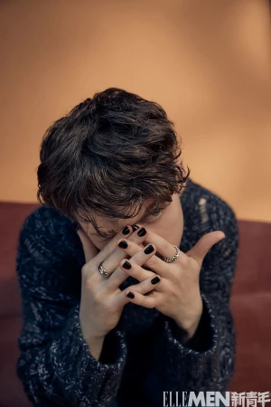Fashion shot of a person with black nail polish and rings, wearing a textured sweater against a warm orange background in a studio.