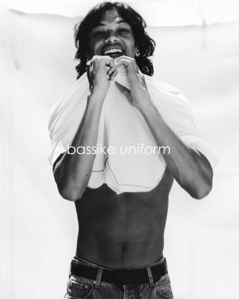 Black and white studio portrait of a smiling adult male, partially undressed, wearing jeans, holding a white t-shirt up to his neck.