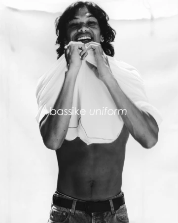 Black and white studio portrait of a smiling adult male, partially undressed, wearing jeans, holding a white t-shirt up to his neck.