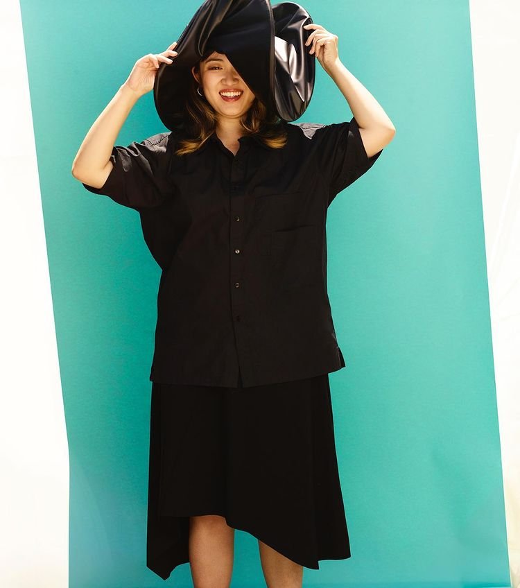 Smiling woman in black button-down shirt and skirt holding up a large curved hat against a blue and white backdrop