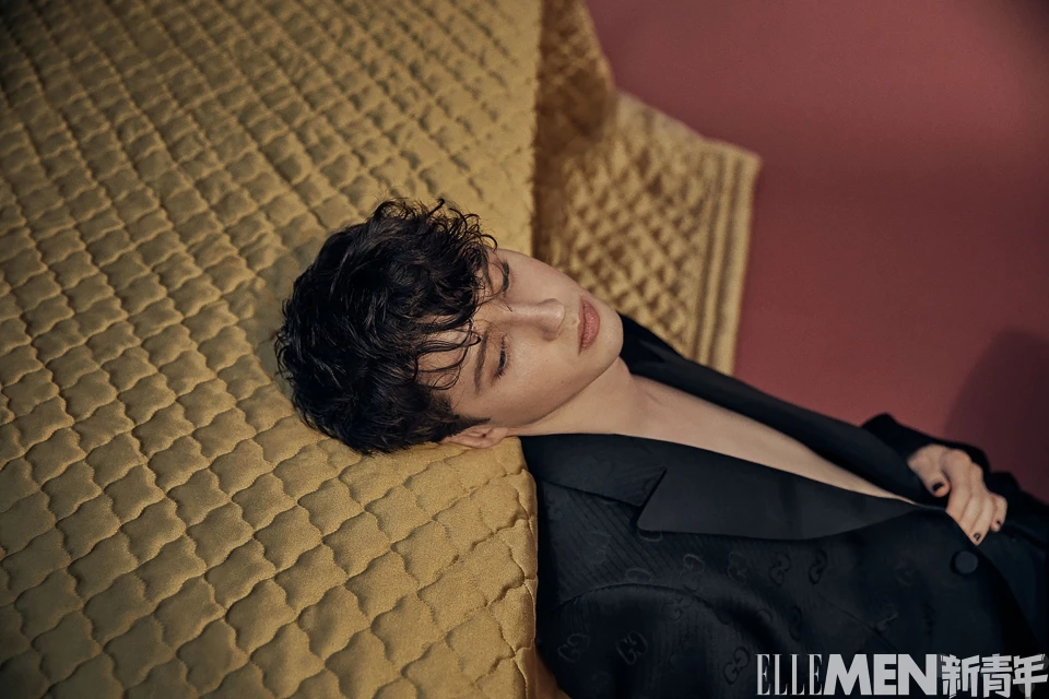 Portrait of a stylish adult male lying on a patterned yellow surface wearing a black blazer against a red and gold background.