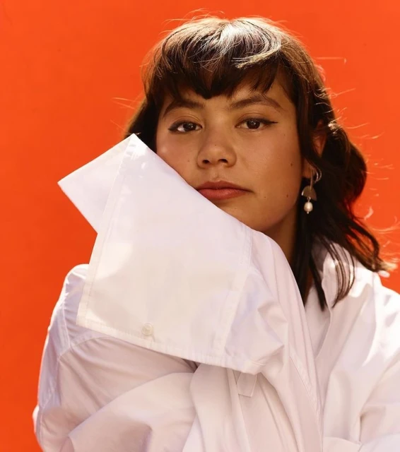 Headshot portrait of a young adult female with dark hair and tan skin, wearing a large white collared shirt against a vibrant orange backdrop.
