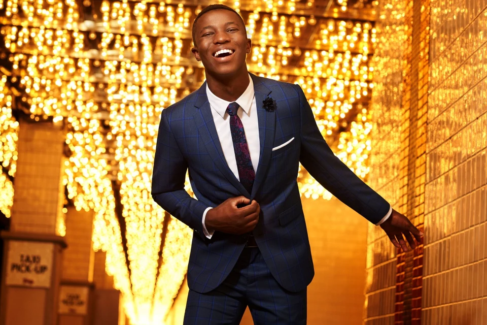 Smiling African American man in a blue suit poses outdoors with warm lighting. Formal wear fashion portrait with celebratory mood.