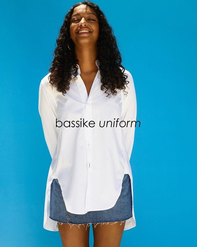 Smiling woman in white button-down shirt and denim shorts poses against a blue background in a medium studio shot. Fashionable and casual.
