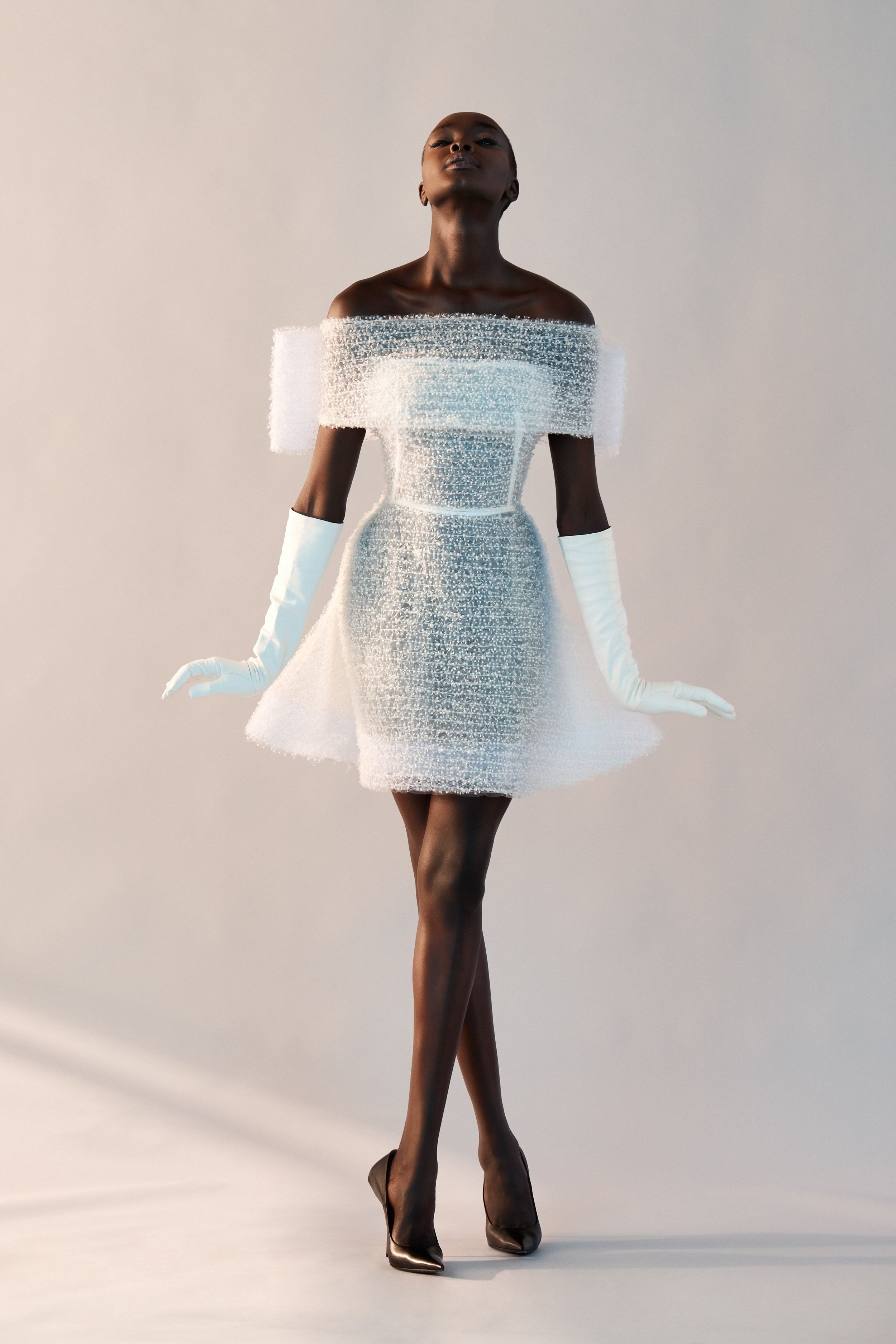 Elegant dark-skinned woman poses in a short, off-the-shoulder white dress with long gloves and high heels against a neutral background.