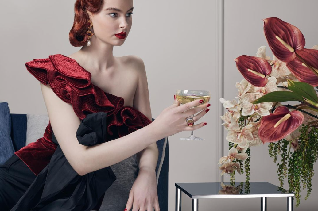 Elegant woman in red dress holds a champagne coupe glass indoors, featuring Anthurium lilies, dark accents, and a light grey backdrop.
