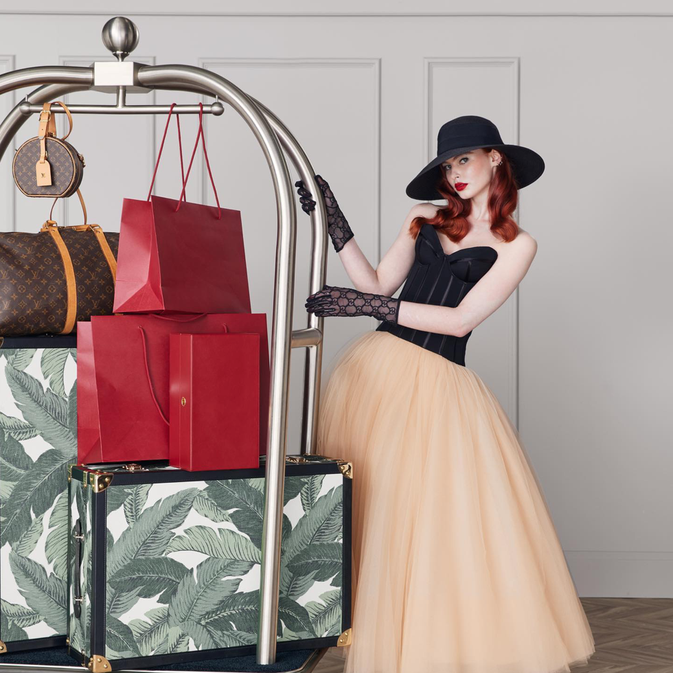 Elegant redhead model in a black corset and tulle skirt poses with vintage luggage and shopping bags on a chrome hotel luggage cart.