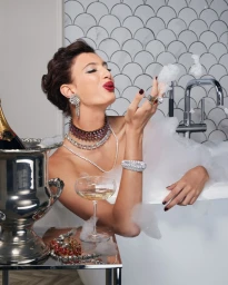 Woman in bathtub with bubbles blowing a kiss wearing layered diamond necklaces and earrings with champagne bucket nearby