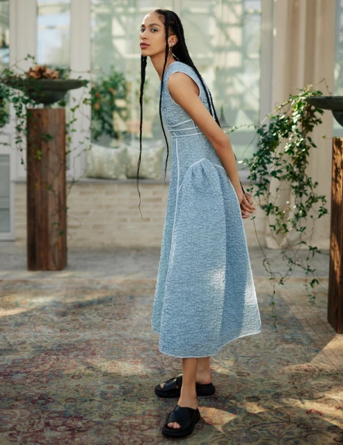 A stylish adult female poses in a light blue textured dress in an indoor garden setting with natural lighting and a vintage carpet.
