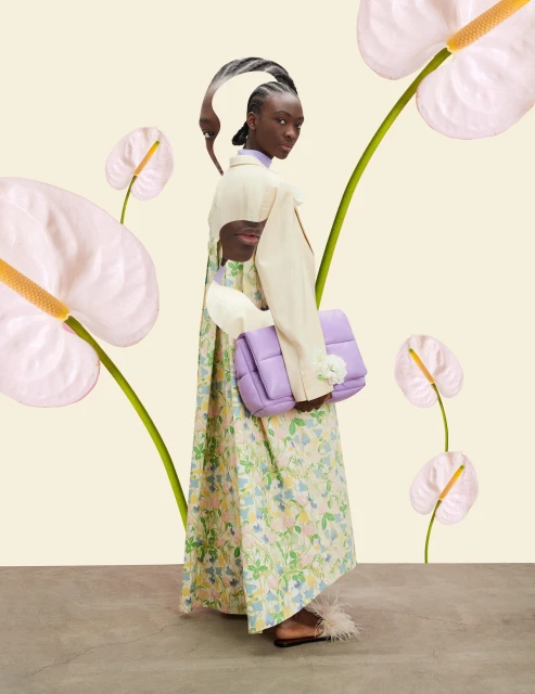 A Black fashion model poses in a dress with a floral pattern while holding a lavender purse, complemented by calla lilies in the background.