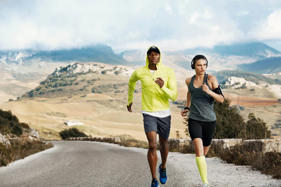 Two athletic adults run on a rural road with scenic mountain backdrop. The man wears yellow jacket and the woman wears a gray tank top.