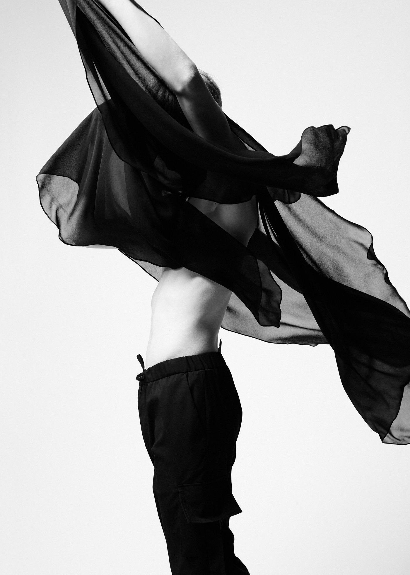 Black and white fashion shot of person with flowing black fabric on a light background, wearing loose pants and revealing midriff.